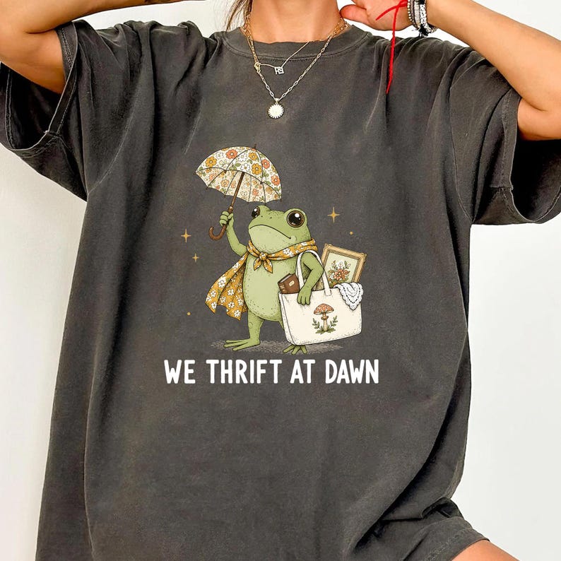 May include: A charcoal gray t-shirt featuring a graphic of a green frog holding a floral umbrella and a white tote bag with a mushroom illustration. The frog wears a floral scarf. Below the graphic, the text reads WE THRIFT AT DAWN in white.