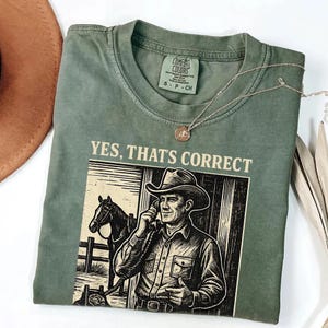 May include: Sage green Comfort Colors t-shirt with the text "YES, THAT'S CORRECT" above a graphic of a cowboy on the phone with a horse in the background. A silver necklace with a pendant is draped across the shirt.