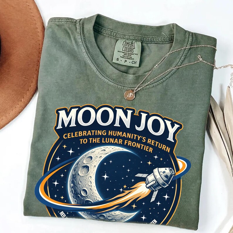 May include: Olive green t-shirt with the text "MOON JOY" and a graphic of the moon, stars, and a rocket ship. The shirt also has the text "CELEBRATING HUMANITY'S RETURN TO THE LUNAR FRONTIER". A silver necklace with a small pendant is on the shirt.