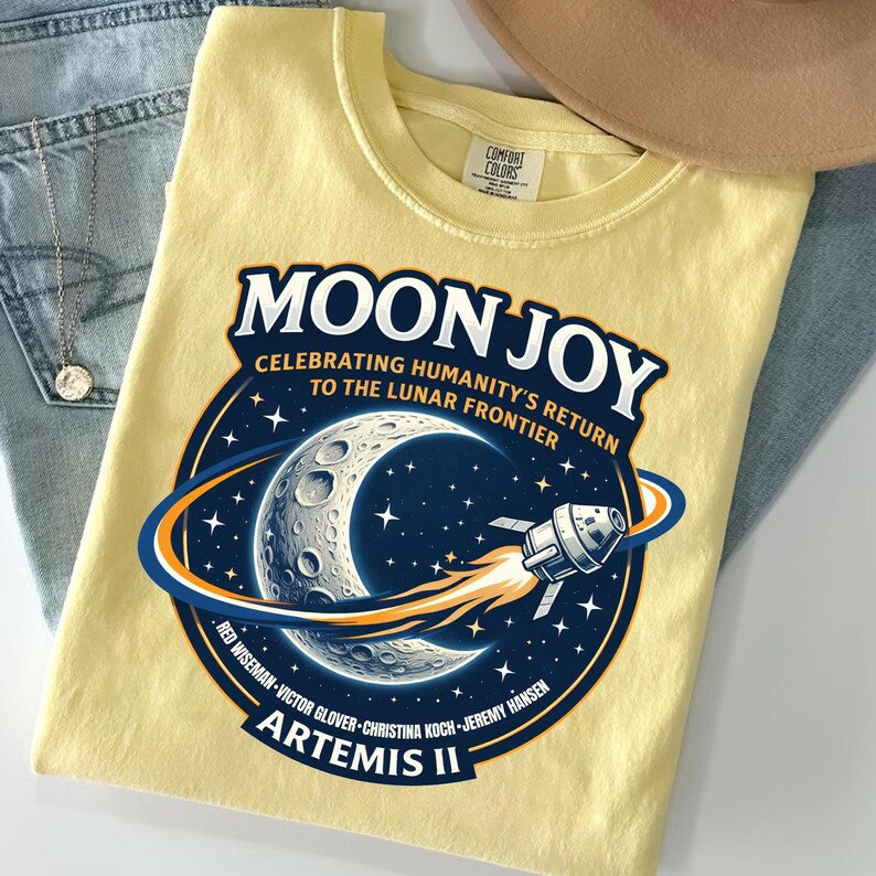 May include: A light yellow t-shirt featuring a circular graphic with the words "MOON JOY" and "CELEBRATING HUMANITY'S RETURN TO THE LUNAR FRONTIER". The graphic includes a crescent moon, stars, and a rocket ship. The shirt also has the text "ARTEMIS II".