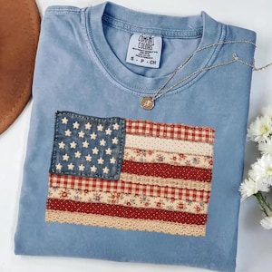 May include: Light blue Comfort Colors t-shirt with a sewn-on American flag design. The flag features a denim blue field with white stars and red and white striped sections made of patterned fabrics. A silver necklace with a pendant rests on the shirt.