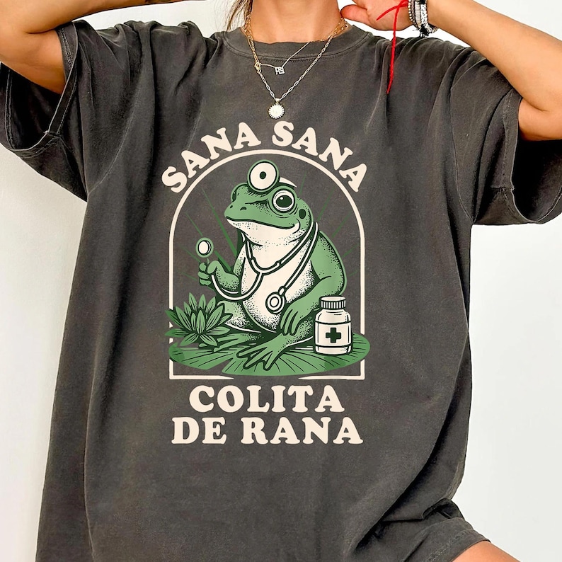 May include: Dark gray t-shirt featuring a green frog doctor with a stethoscope and a medicine bottle. The shirt has the words "Sana Sana" above the frog and "Colita de Rana" below it, all in white lettering.