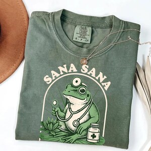 May include: Green Comfort Colors t-shirt with a graphic of a frog doctor wearing a stethoscope and holding a medical tool. The shirt has the words "Sana Sana" in a circular design. A silver necklace and a brown hat are also visible.