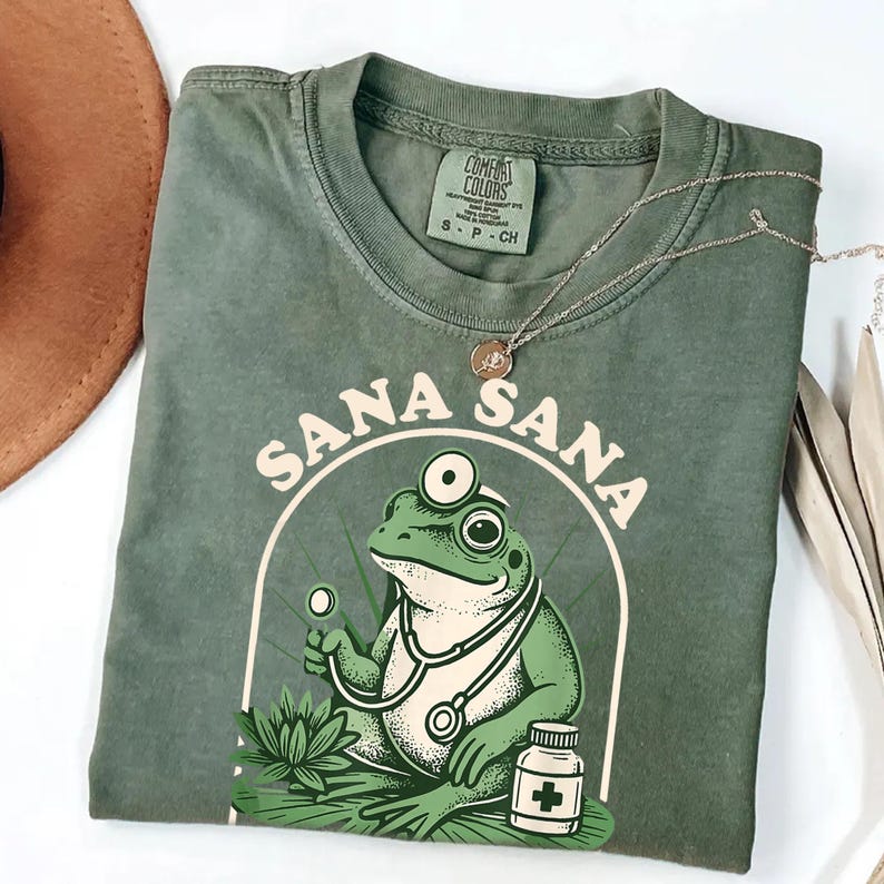May include: Green Comfort Colors t-shirt with a graphic of a frog doctor wearing a stethoscope and holding a medical tool. The shirt has the words "Sana Sana" in a circular design. A silver necklace and a brown hat are also visible.