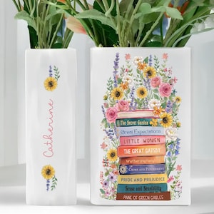 May include: Two white ceramic vases. One vase has the name "Catherine" in pink script with sunflower and floral accents. The other vase features a stack of classic book titles like "Pride and Prejudice" and "The Great Gatsby" surrounded by flowers.