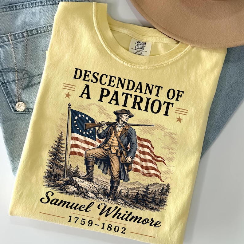 May include: A light yellow t-shirt featuring a graphic of a man in colonial attire holding a rifle, with an American flag in the background. The text reads "Descendant of a Patriot" and "Samuel Whitmore 1759-1802". The shirt is folded on a pair of blue jeans.