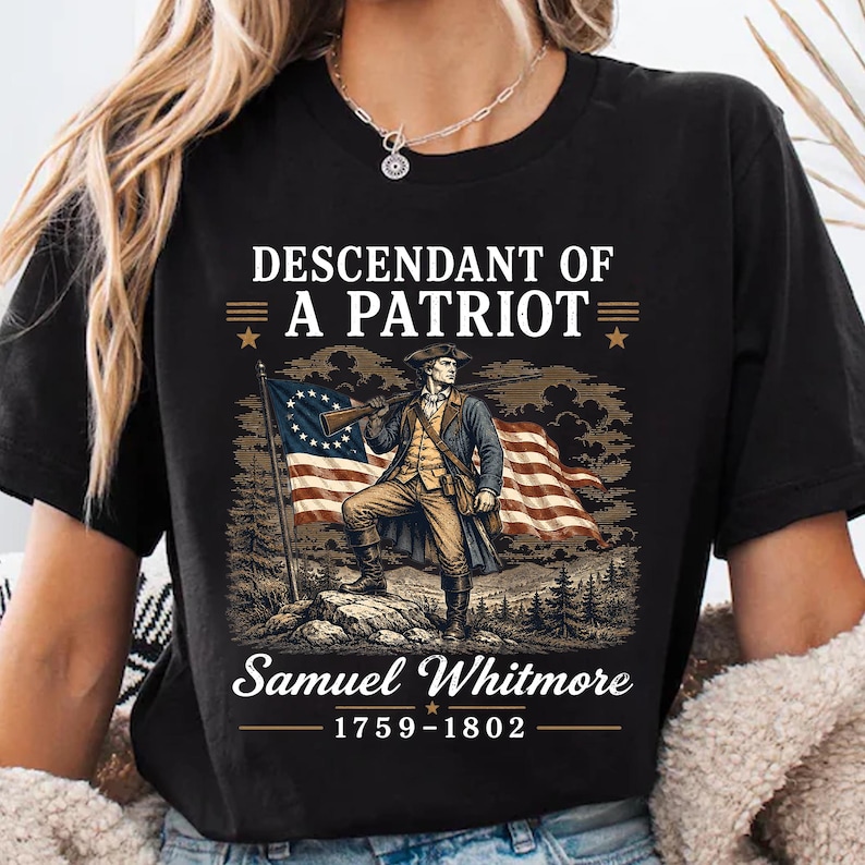 May include: Black t-shirt featuring a patriotic graphic. The design includes an illustration of a man in historical attire holding a rifle, standing before a flag. Text reads "Descendant of a Patriot" and "Samuel Whitmore 1759-1802".
