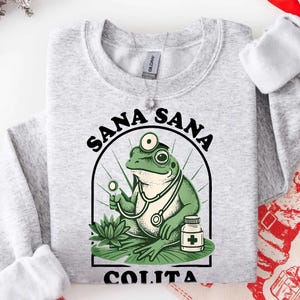 May include: A light gray sweatshirt featuring a green frog doctor with a stethoscope, holding a medical tool, and a medicine bottle. The text "SANA SANA COLITA" is printed above and below the frog illustration.