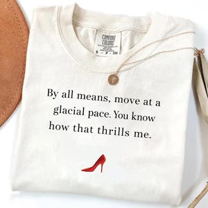 May include: Off-white t-shirt featuring the quote "By all means, move at a glacial pace. You know how that thrills me." and a red high heel illustration. A gold necklace with a circular pendant rests on the shirt.