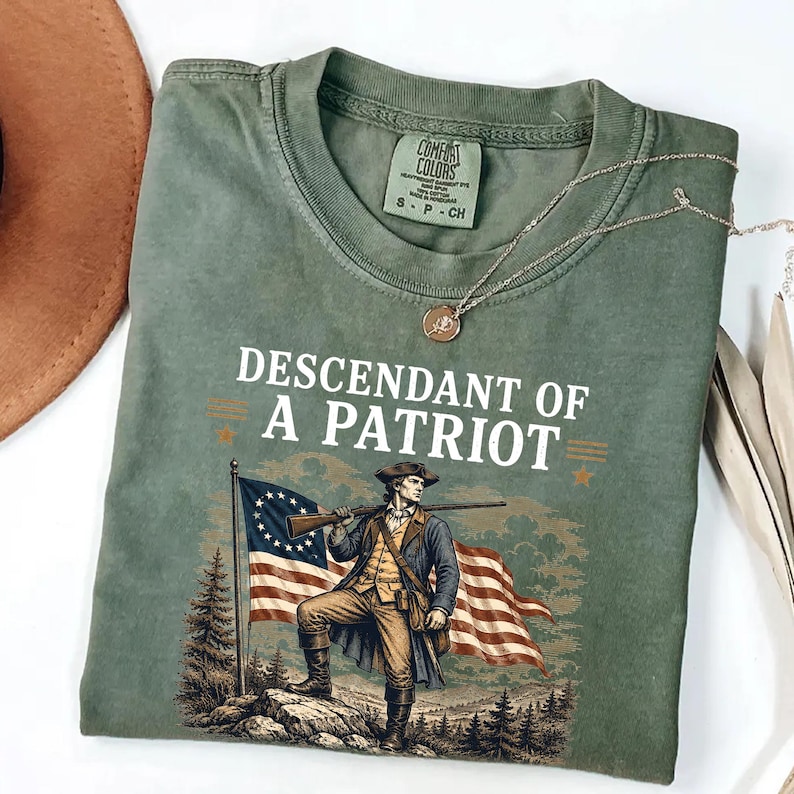 May include: Olive green Comfort Colors t-shirt with the text "DESCENDANT OF A PATRIOT" above an illustration of a colonial soldier holding a rifle and standing in front of an American flag. A gold necklace is draped across the shirt.