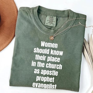 May include: An olive green t-shirt with the text "Women should know their place in the church as apostle prophet evangelist" in white. A gold necklace with a pendant is on the shirt. The shirt is folded and next to a brown hat.