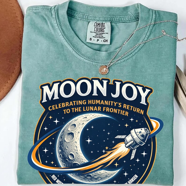 May include: A seafoam green t-shirt with the words "MOON JOY" and a graphic of the moon, stars, and a spaceship. The shirt also has the text "CELEBRATING HUMANITY'S RETURN TO THE LUNAR FRONTIER". A silver necklace with a pendant rests on the shirt.