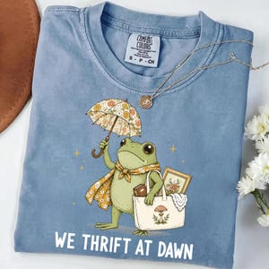 May include: A blue short-sleeve t-shirt featuring a graphic of a green frog holding a floral umbrella and a tote bag containing a book and a framed picture. The text WE THRIFT AT DAWN is printed below the frog. The shirt has a Comfort Colors label.