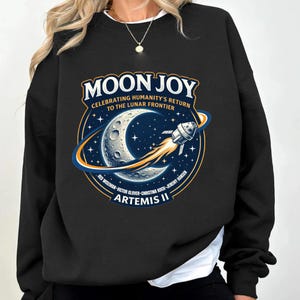 May include: Black sweatshirt featuring a graphic with the text "MOON JOY" and "CELEBRATING HUMANITY'S RETURN TO THE LUNAR FRONTIER." The design includes a crescent moon, stars, and a rocket ship. The names of the Artemis II crew are listed below.