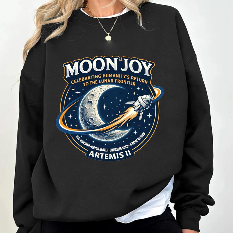 May include: Black sweatshirt featuring a graphic with the text "MOON JOY" and "CELEBRATING HUMANITY'S RETURN TO THE LUNAR FRONTIER." The design includes a crescent moon, stars, and a rocket ship. The names of the Artemis II crew are listed below.