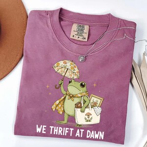 May include: A mauve short-sleeved t-shirt featuring a graphic of a green frog holding a floral umbrella and a tote bag. The frog wears a floral scarf. The text WE THRIFT AT DAWN is printed in white below the frog. The shirt has a Comfort Colors label.