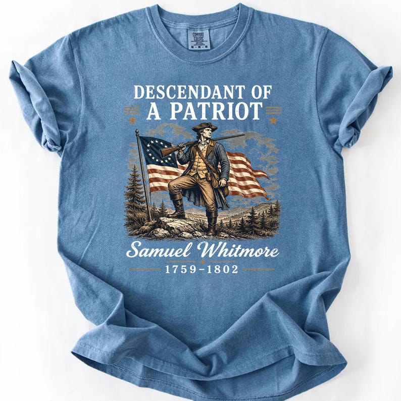 May include: Blue t-shirt featuring a graphic of a man holding a rifle in front of an American flag. The text reads "Descendant of a Patriot" and "Samuel Whitmore 1759-1802".