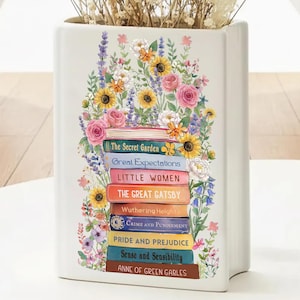 May include: White ceramic vase shaped like a book, decorated with a stack of classic literature titles and floral arrangements. Titles include "The Secret Garden," "Great Expectations," and "Pride and Prejudice." The vase is adorned with sunflowers, roses, and other flowers.
