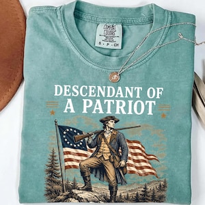 May include: A seafoam green t-shirt with the words "DESCENDANT OF A PATRIOT" above an illustration of a man holding a rifle and standing in front of a flag. A silver necklace with a pendant is draped over the shirt.