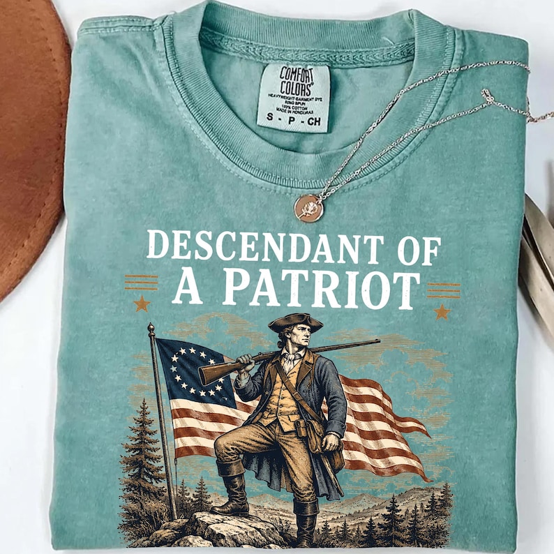 May include: A seafoam green t-shirt with the words "DESCENDANT OF A PATRIOT" above an illustration of a man holding a rifle and standing in front of a flag. A silver necklace with a pendant is draped over the shirt.