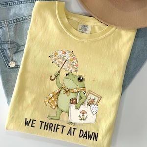May include: A pale yellow t-shirt featuring a graphic of a green frog holding a floral umbrella and carrying a tote bag containing a framed picture and a book. The text "WE THRIFT AT DAWN" is printed in black below the frog illustration.