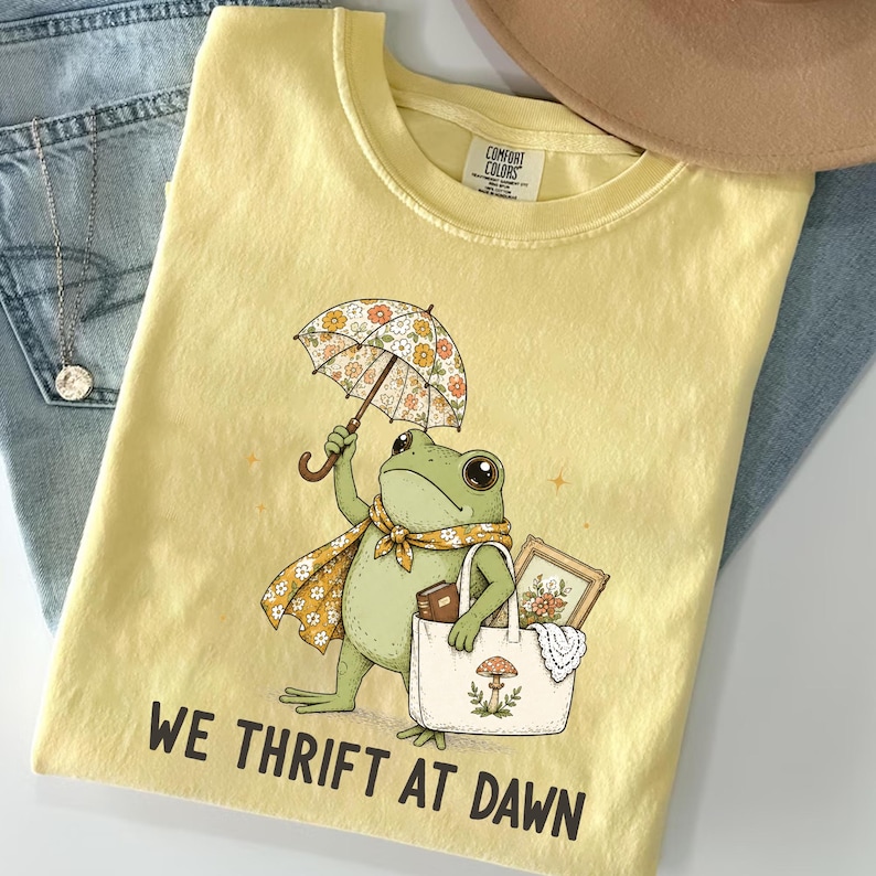 May include: A pale yellow t-shirt featuring a graphic of a green frog holding a floral umbrella and carrying a tote bag containing a framed picture and a book. The text "WE THRIFT AT DAWN" is printed in black below the frog illustration.