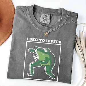 以下が含まれることがあります： 「I BEG TO DIFFER」の文字とナイフを持った緑色のカエルのイラストがプリントされたグレーのComfort Colors Tシャツ。小さなペンダントが付いたシルバーのネックレスがシャツの上にかけられています。シャツは白い表面に折りたたまれています。