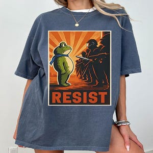 May include: A blue t-shirt with a graphic depicting a cartoon frog in a blue scarf confronting silhouetted figures with batons. The word "RESIST" is printed in bold letters at the bottom of the graphic, in a retro style.