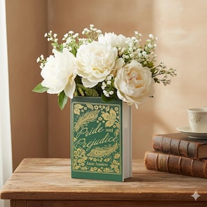 May include: A rectangular, green vase shaped like a book, featuring the title "Pride and Prejudice" in gold lettering, holds a bouquet of white peonies and small white flowers. The vase sits on a wooden surface, with a teacup and books in the background.