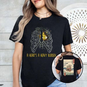 Op de afbeelding: Zwart t-shirt met een afbeelding van een ribbenkast met een geel hart en de tekst "A HEART'S A HEAVY BURDEN". De achterkant van het shirt heeft een illustratie in Japanse stijl.