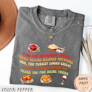 May include: Pepper-colored Comfort Colors t-shirt with a Thanksgiving-themed graphic. The graphic includes illustrations of cranberry sauce, mashed potatoes, a turkey, rolls, and a glass of wine, with text that reads "Pass the Cranberry Sauce".