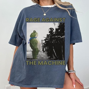 May include: Blue-grey t-shirt with the text "RAGE AGAINST THE MACHINE." The graphic shows a green frog with a blue scarf confronting figures in black uniforms. The shirt has short sleeves and a relaxed fit.