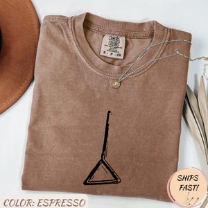 May include: Espresso-colored Comfort Colors t-shirt with a black graphic design. A silver necklace with a pendant rests on the shirt. The shirt has a crew neck and short sleeves. The text "SHIPS FAST!" is in a pink circle.