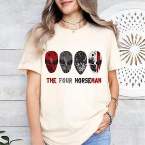 May include: Cream-colored t-shirt featuring four stylized masks in red, gray, and black. The text "THE FOUR HORSEMAN" is printed in red below the masks. The shirt is a casual wear item.
