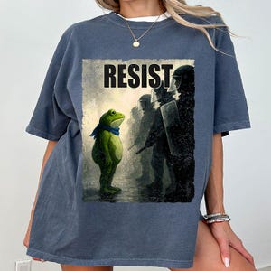 Puede incluir: Camiseta azul con la palabra "RESIST" sobre una ilustración de una rana frente a una línea de antidisturbios. La rana lleva una bufanda azul. La imagen tiene un estilo artístico y desgastado.
