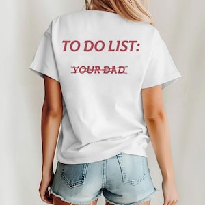 May include: White t-shirt with the text "TO DO LIST: YOUR DAD" in red. The words "YOUR DAD" are crossed out. The shirt is worn by a person wearing denim shorts.