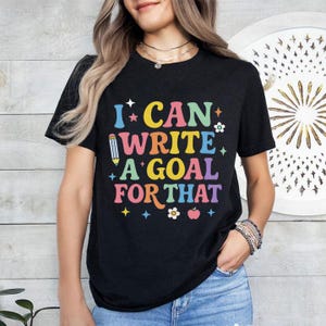 Könnte beinhalten: Schwarzes T-Shirt mit dem farbenfrohen Schriftzug "I CAN WRITE A GOAL FOR THAT". Das Design beinhaltet einen Bleistift, Sterne, Blumen und einen Apfel. Das T-Shirt hat einen Rundhalsausschnitt.