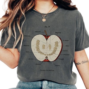 May include: Dark grey t-shirt featuring a detailed illustration of an apple cut in half, showcasing the anatomy of teeth and labeled parts. The text "fig. 34" and "Dentes Os Malum" are also visible on the shirt.