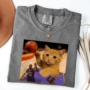 May include: Gray Comfort Colors t-shirt featuring a basketball-themed graphic. The graphic shows an orange tabby cat playing basketball with a basketball and a basketball player. A silver necklace with a pendant is on the shirt.