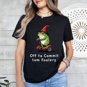 Puede incluir: Camiseta negra con una rana verde con un sombrero de bruja rojo montada en un scooter rojo y negro. El texto "Off to Commit tom Foolery" está impreso debajo.