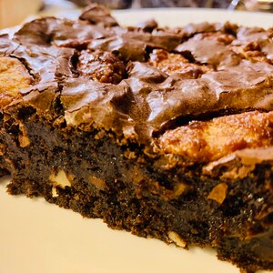 May include: A close-up of a chocolate brownie with a fudgy texture and a rich, dark brown color. The brownie is topped with pieces of caramel and nuts.