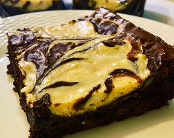 May include: A close-up of a chocolate brownie with a swirl of vanilla cheesecake on top. The brownie is on a white plate.