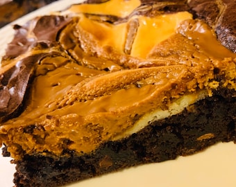 May include: A close-up of a chocolate brownie with a swirl of caramel on top. The brownie is cut into a slice and is sitting on a white plate.