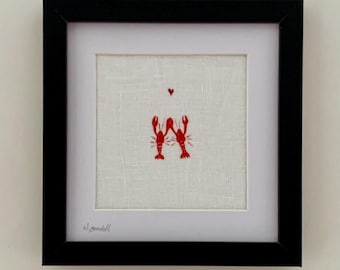 May include: A square black frame containing a white fabric mat and a central embroidery on off-white linen. The embroidery features two red lobsters with their claws touching and a small red heart above them. The artist signature is in the corner.
