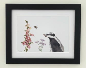 May include: A framed watercolour print showing a badger, a bee, and flowers. The badger is in black and white, with a pink and purple flower and a bee in flight. The print is framed in a black frame.