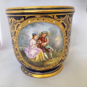 May include: A blue and gold porcelain planter with a painted scene of a couple in a garden. The planter has a rounded shape with a gold rim and base, and is decorated with gold filigree and small jewels.