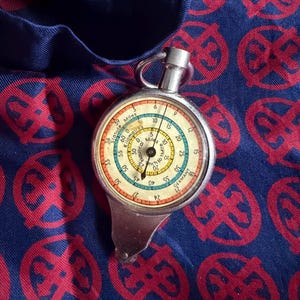 May include: A vintage silver map measurer with a circular dial showing miles and kilometres. The dial has a cream background with black numerals and red, blue, and yellow borders. The measurer sits on navy blue fabric with a red pattern. The dial measures in inches.