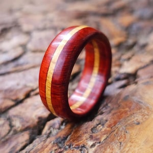 May include: A wooden ring with a reddish-brown exterior and a light-colored wood stripe running through the center. The ring is made of two different types of wood and has a polished finish. The ring is a jewelry item.