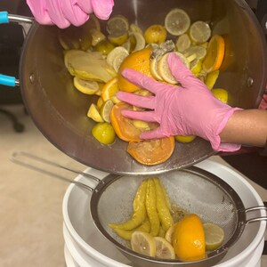 Pu&ograve; includere: Una persona con guanti rosa versa una miscela di fette di limoni, lime e arance da una ciotola di acciaio inossidabile in uno scolapasta sopra una ciotola bianca.