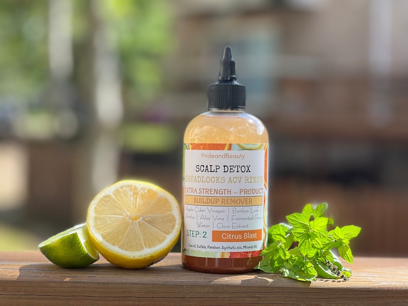 Pu&ograve; includere: Una bottiglia di plastica trasparente con un tappo nero e un'etichetta gialla. L'etichetta riporta "PrideandBeauty Scalp Detox Dreadlocks ACV Rinse Extra Strength - Product Build Up Remover Apple Cider Vinegar | Bamboo Leaf | Aloe Vera | Fermented Rice Water | Clove Extract Step: 2 Citrus Blast". La bottiglia &egrave; appoggiata su una superficie di legno con un limone e un lime.